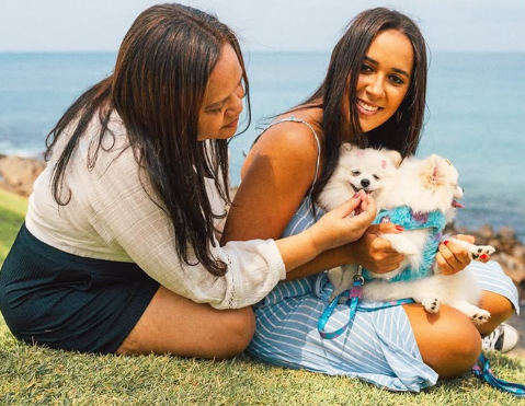 Duas tutoras sentadas na grama à beira-mar com um Pomeranian branco no colo